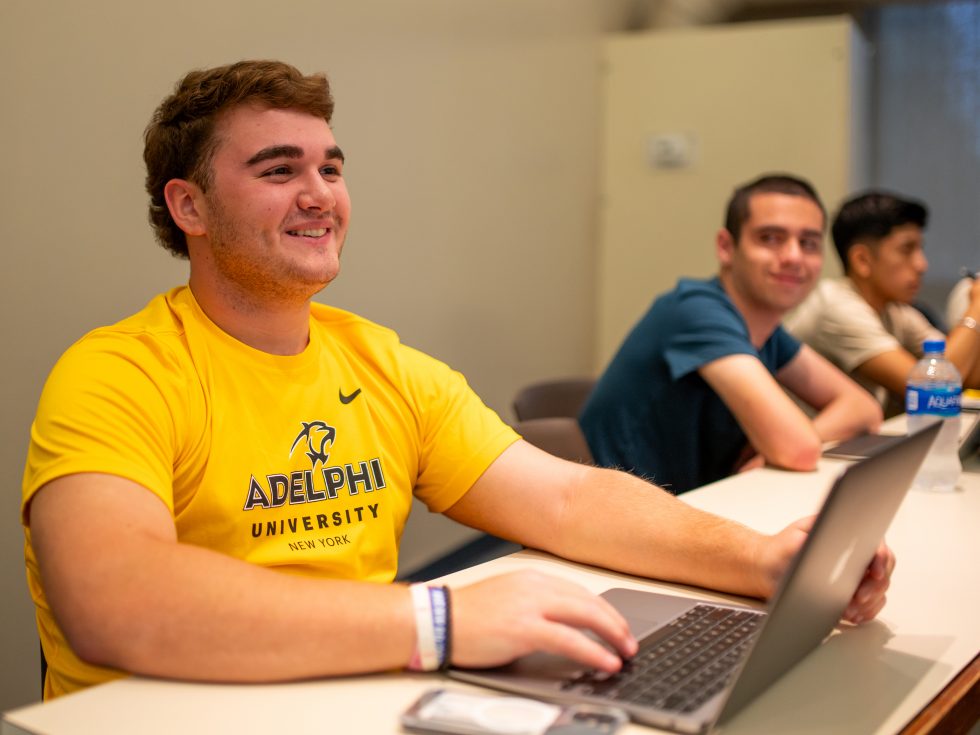 Smiling male student wearing a gold Adelphi shirt in class.
