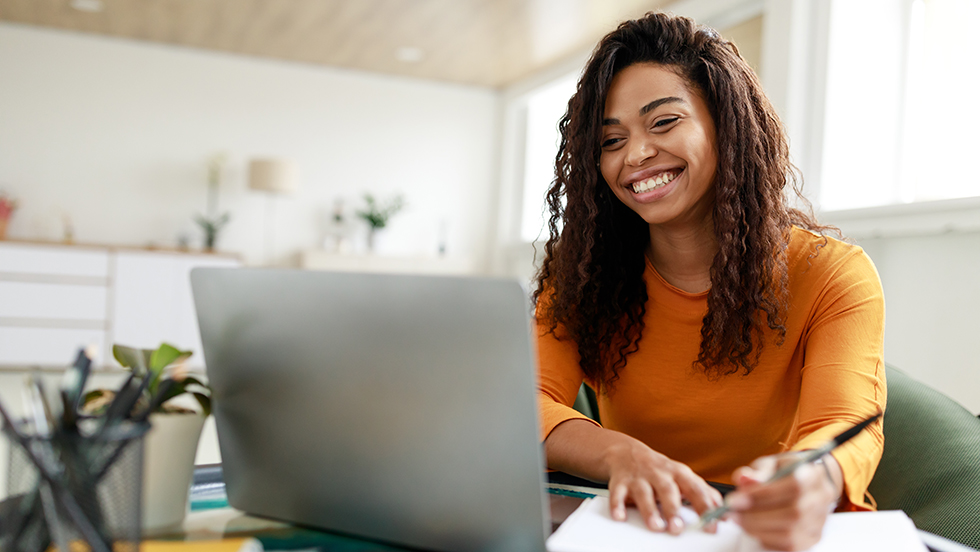 A young woman works at home on a laptop computer while taking notes by hand in a notebook.