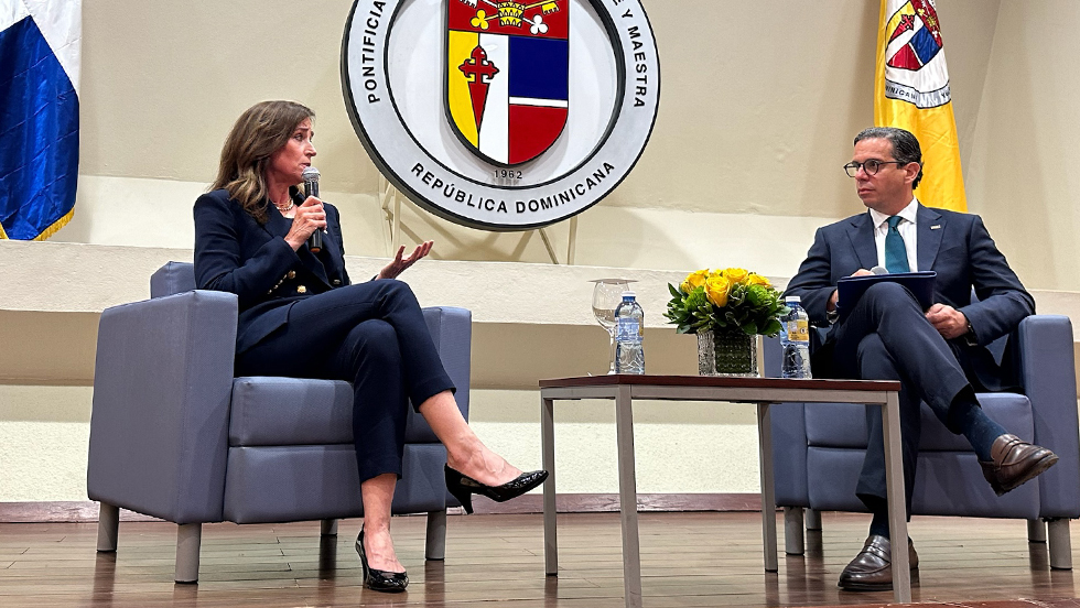 Woman in blue suit in blue chair on stage, holding microphone, speaking to a man in a suit seated next to her. Emblem displayed in background reads "Pontificia Universidad Católica Madre y Maestra"