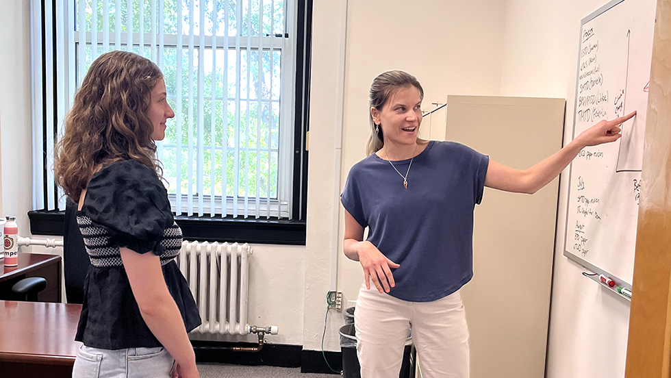 Woman in blue shirt points to white board on the right, woman in black shirt faces white board.