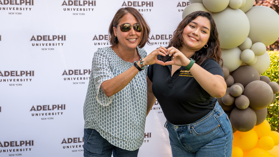 Two women stand side by side and create a heart symbol with their hands. The backdrop behind them reads "Adelphi University," and there are yellow, black and gray balloons to the right.