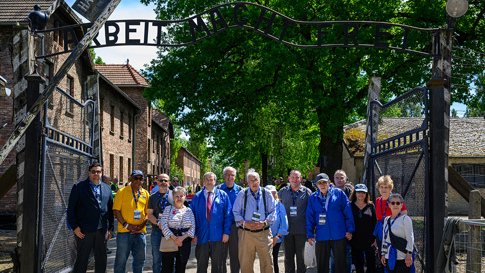 The Dr. Machlis and the group of education leaders and their spouses pose under the "Arbeit Macht Frei" sign over the entrance at Auschwitz. 