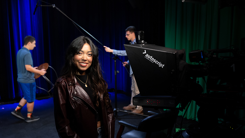 A young woman sets up production equipment in a television station, as two men behind her setting up a microphone and seating.