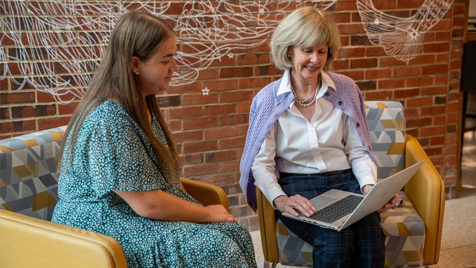 Two women sitting on chairs, one on the right holds a laptop that they both look at.