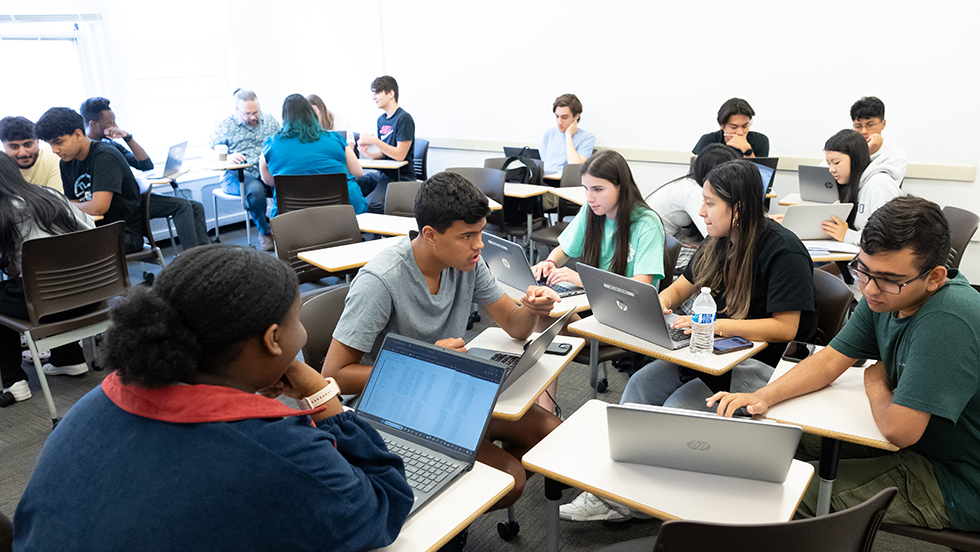 Students work together in groups in a classroom setting, with laptop computers on their desks.