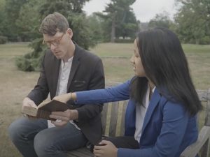 Man sits on left with open book, young woman sits next to him pointing at the open page