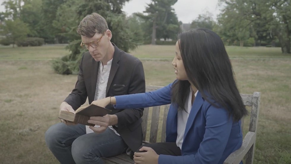 Man sits on left with open book, young woman sits next to him pointing at the open page