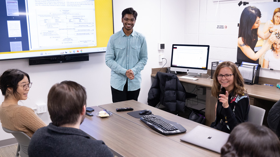 A male college student stands in front of a table where three people are seated. A laptop, keyboard and mouse are on the table. The woman seated is pointing up and speaking. Behind the student is a whiteboard with a chart. In the background to the right is a poster of people putting an electrode cap on a woman to do neurological research.