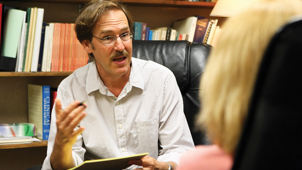 A man seated in front of a bookshelf is talking animatedly.