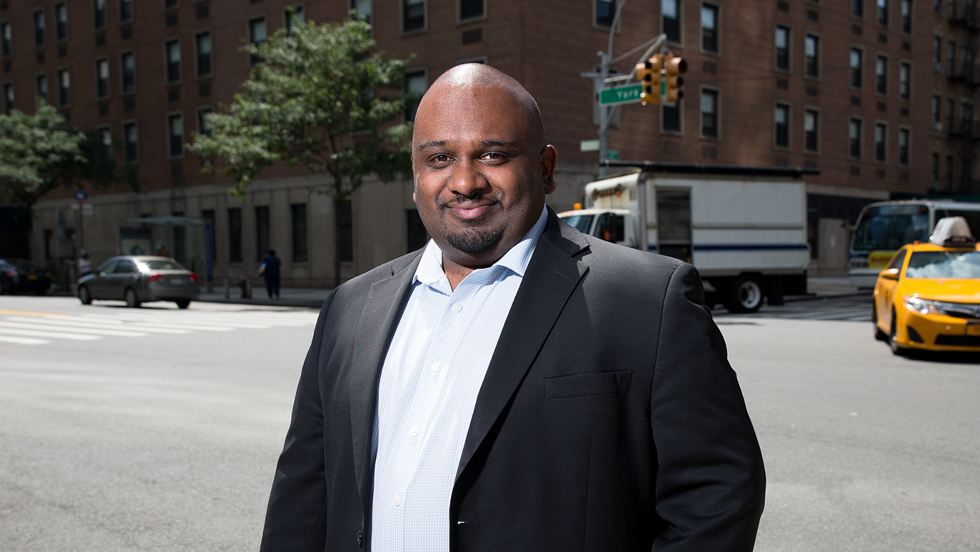 Headshot of a man outside in a button-up shirt and blazer, smiling at camera