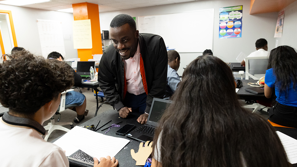 MBA student Mujjabi Ian Sebbowa leans in to assist a classroom full of teens, seated at their computers.