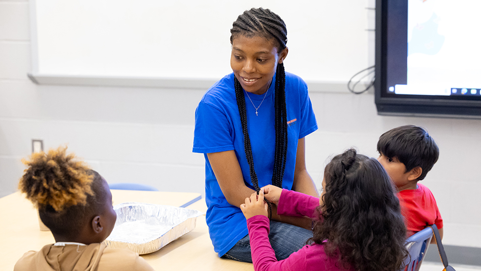 Biology Graduate Student Peace Olutoye, perched on a desk, sits with several children in a classroom setting, smiling.