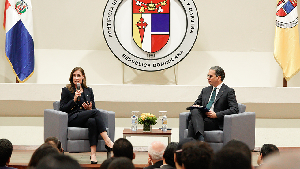 President Riordan is seated in front of a large university seal, holding a microphone. To her right is a professor in that university. 