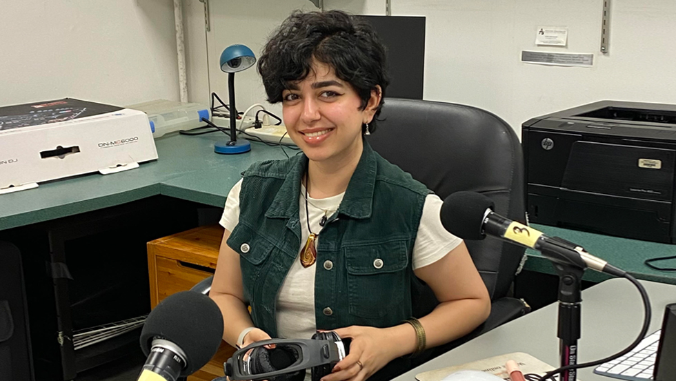 A young woman sits at a desk in front of a microphone. She holds headphones.