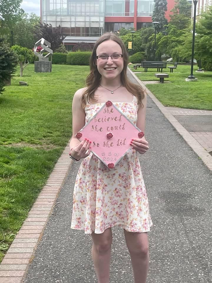 Young woman holding up decorated graduation cap, smiling at camera. The cap says: "She believed she could, so she did."