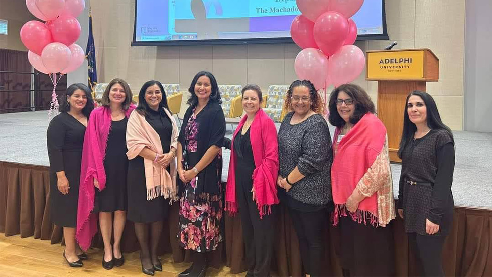  Eight women accessorized in pink standing in front of stage flanked by pink balloons.