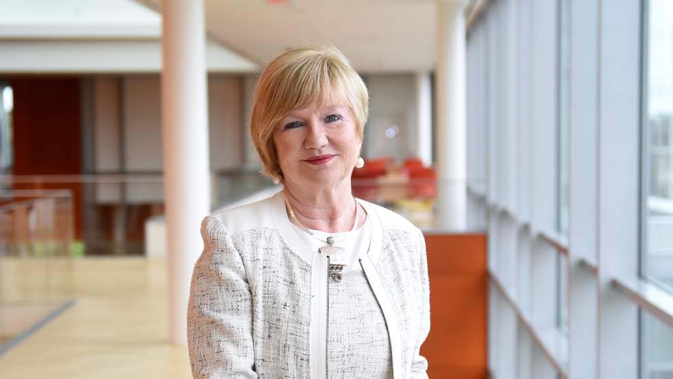 A woman in white smiling. In the background is the lobby of Adelphi University’s Nexus Building.