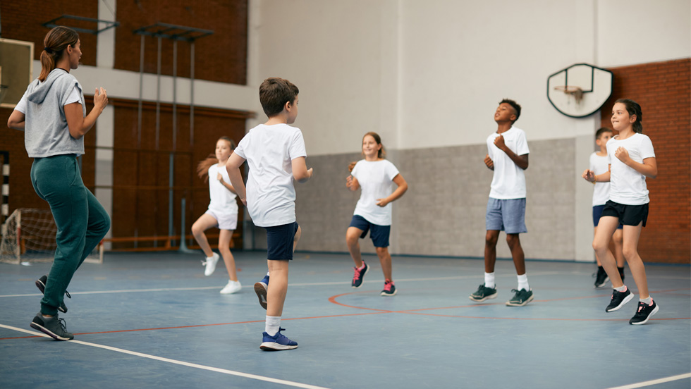 A female instructor leads six young students through exercises in a school gym class.