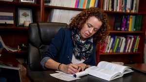 Professor Fiori at her desk, writing on a pad of paper that sits next to an open book.