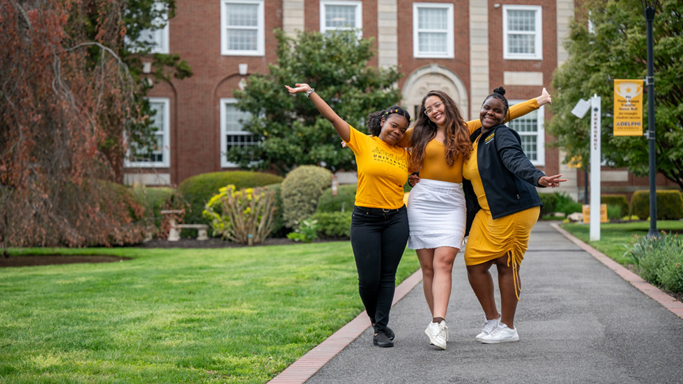 Three women students stand in pathway, arms raised triumphantly, on pathway in front of Adelphi’s Levermore Hall. Each wears Adelphi’s signature color, gold.