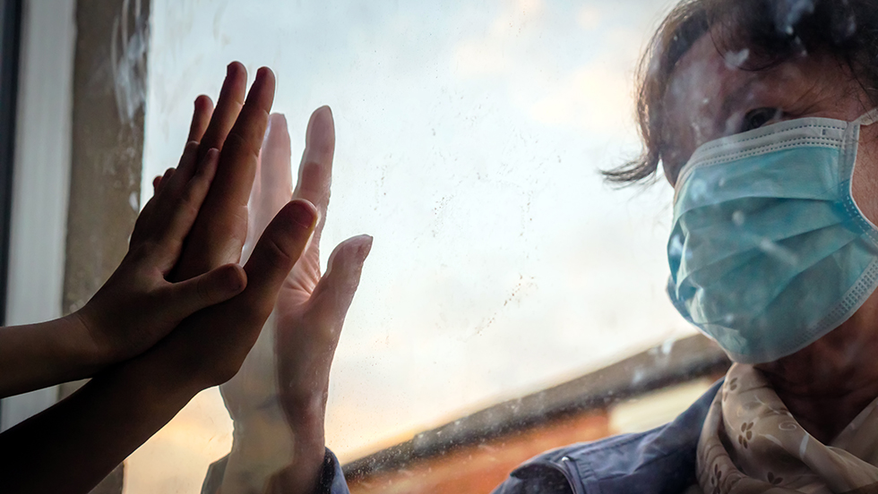 A woman wearing a surgical mask puts her hand to a window opposite the hands of another adult and a child.