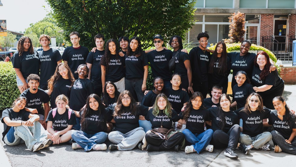 29 students in three rows outdoors, face the camera. They wear shirts that read “Adelphi Gives back.”