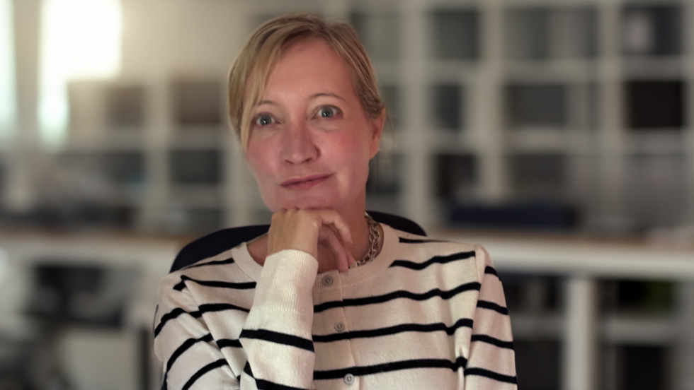 A blonde woman wearing a black-and-white striped blouse looks ahead, her chin resting on her right hand