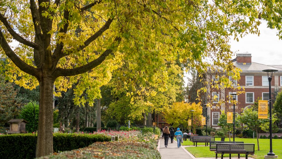 A few students walking on a path toward a brick building on Adelphi’s campus with trees, flowers and tables along the side 