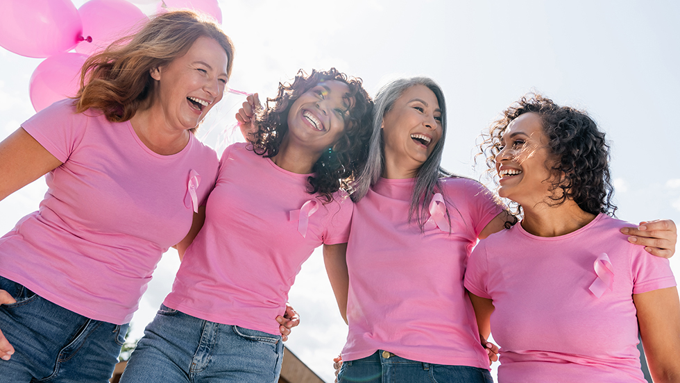 A group of women smile with arms around one another. They wear pink shirts with the breast cancer pink ribbon pins.