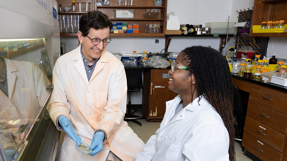 Dr. Heyl and Kyana Gordon in his lab. Both wear lab coats. Dr. Heyl has a lab container holding algae in his hands.