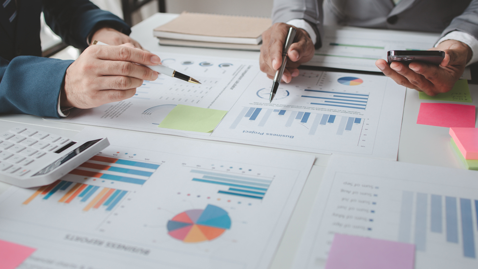 Two men, whose hands are only seen, at a table covered by documents with charts and graphs.