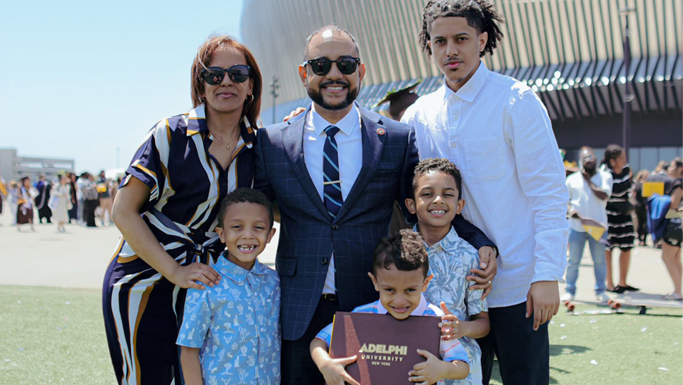 Center, a man in suit with sunglasses. A woman in sunglasses stands to his right. Surrounding them are four boys, one of whom holds a diploma case that reads “Adelphi University New York.”