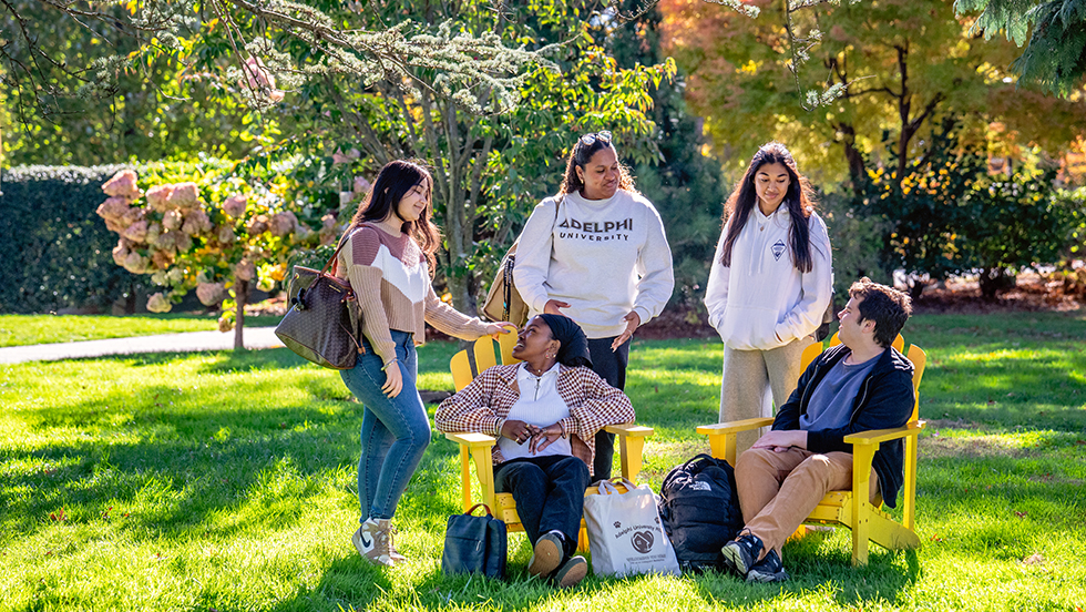 A group of students gathered on a campus lawn, interacting. The young woman in the center wears a sweatshirt that reads "Adelphi University"