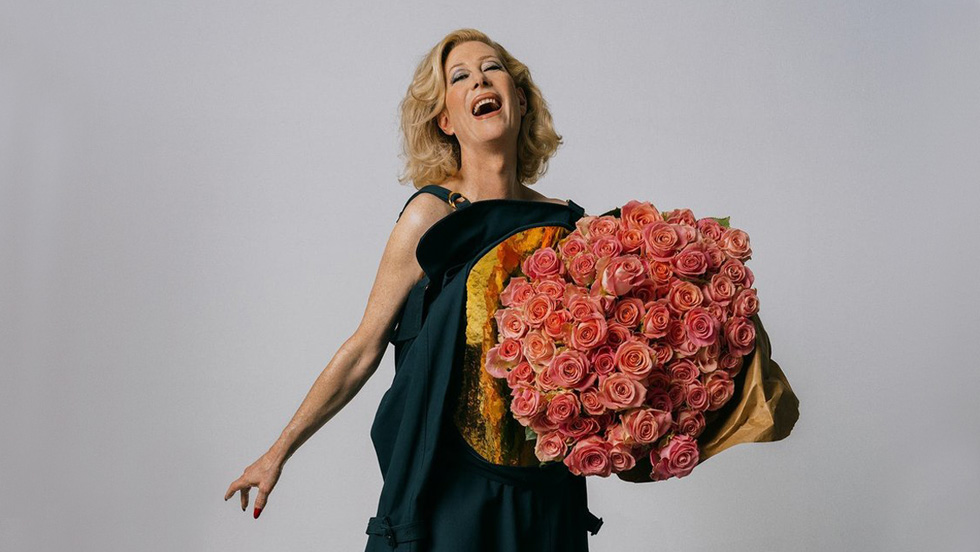 A nonbinary person with shoulder-length blond hair wearing a black dress smiles widely while holding a huge bouquet of roses.