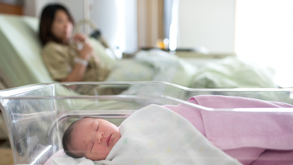 A newborn in a bassinet inside a hospital room, with her mother in the background in a hospital bed.