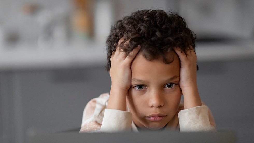 A young boy stares ahead, looking at a computer screen. He has his hands on the sides of forehead, in a pose of concentration.