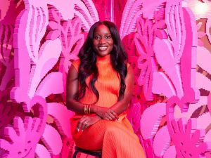 A young Black woman in an orange dress sits in front of a pink Lego sculpture.