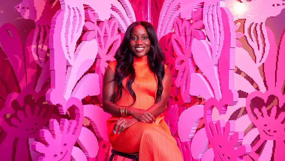 A young Black woman in an orange dress sits in front of a pink Lego sculpture.