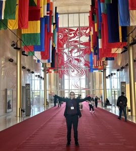 A man, arms outstretched, standing beneath flags in a hallway