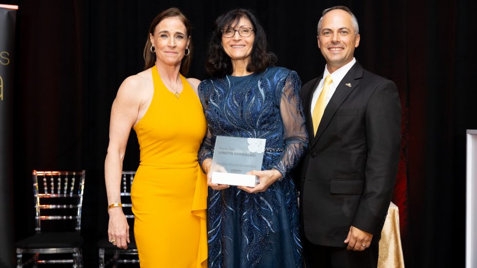 From left to right: a woman in a yellow dress, a woman in a blue embroidered gown holding an award plaque, and a man in a black suit with a yellow tie.