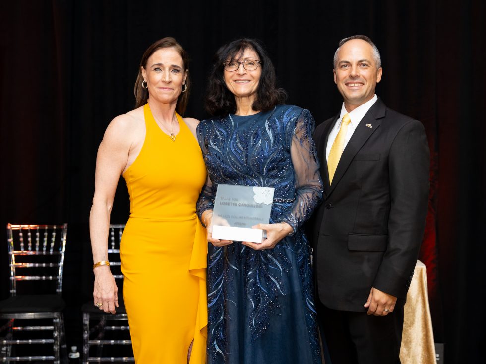 From left to right: a woman in a yellow dress, a woman in a blue embroidered gown holding an award plaque, and a man in a black suit with a yellow tie.