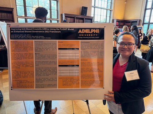 A young woman with dark hair pulled back and wearing eyeglasses stands, smiling, before her poster presentation in a bright auditorium. 