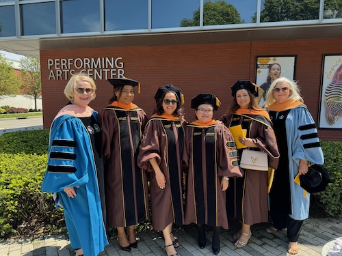 Four young women in their doctoral robes are flanked by two female faculty members, both with light blonde hair and sunglasses. They are standing in front of a Performing Arts Center. 