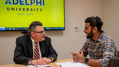 Thomas Ward and Husein Ghadiali in discussion at a table in the career center. A television screen behind them shows the Adelphi University logo.