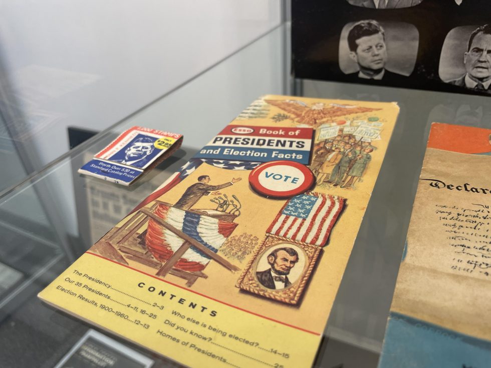 A collection of election memorabilia on a glass case, including a book of stamps with the image of John F. Kennedy, a “Book of Presidents and Election Facts,” photos of John F. Kennedy and Richard Nixon, and a partially visible pamphlet of the Declaration of Independence.