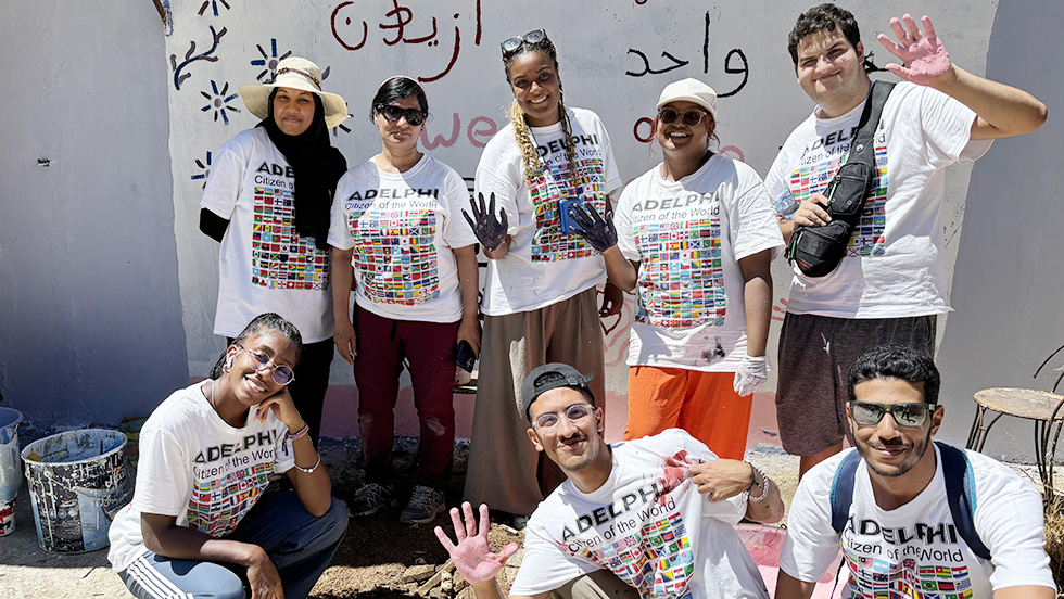 Eight students standing in front of a mural they have painted on the side of a school building. The students are wearing t-shirts that say "Adelphi Citizen of the World" and are showing their paint-covered hands.