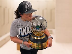 Heather Mau in the team's training room kissing the championship trophy, which she holds in her arms.  The trophy is a silver basketball on a wood pedestal with gold plating.  She is wearing a t-shirt and hat given the players and training staff commemorating the Western Conference championship.