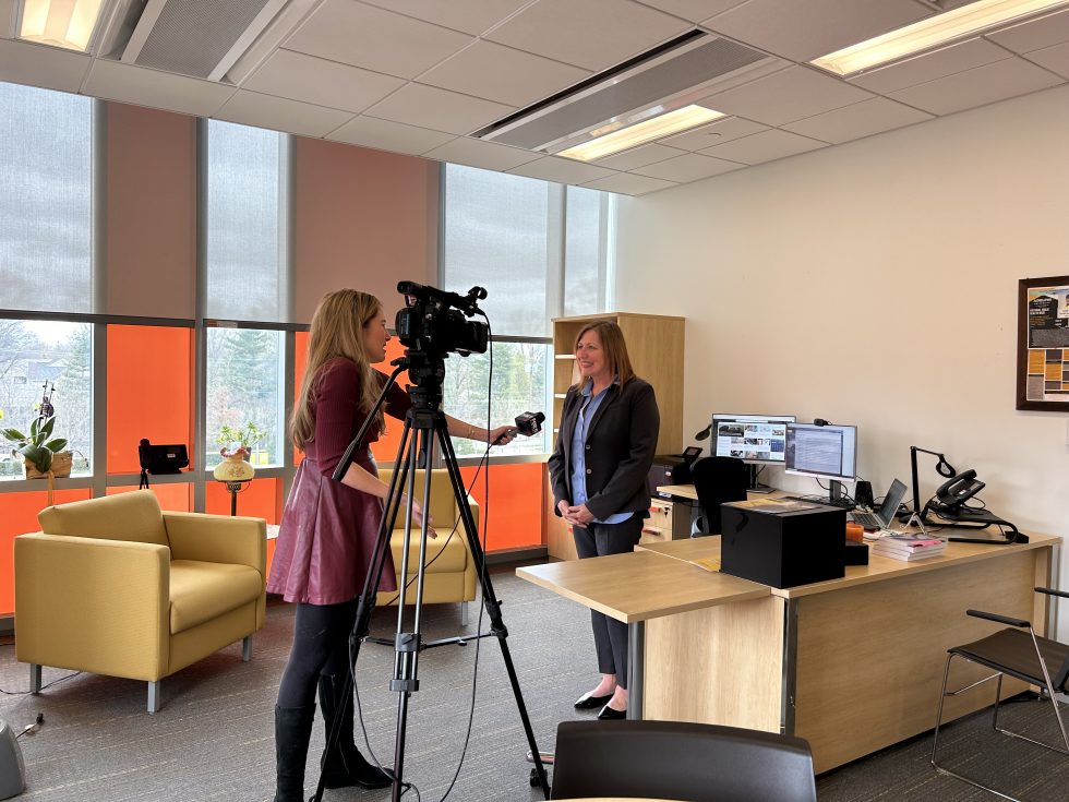 A female reporter with a camera and microphone interviewing Adelphi’s Dean of the college of nursing and public health Deborah Hunt in her office.