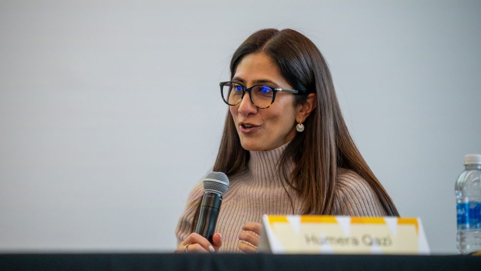 A woman seated at table holding a microphone and speaking. The name card in front of her reads "Humera Qazi."