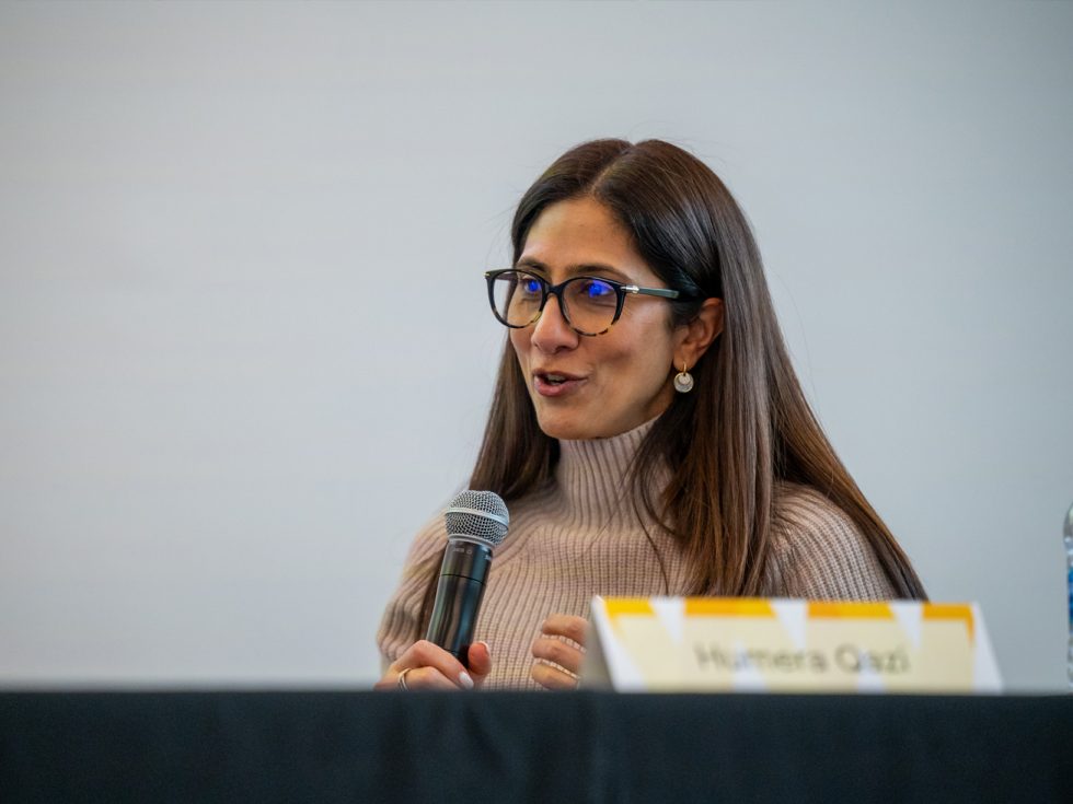 A woman seated at table holding a microphone and speaking. The name card in front of her reads "Humera Qazi."